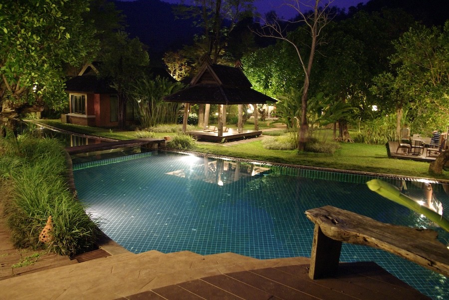  A backyard oasis at night with a glimmering pool and a pergola illuminated by outdoor lighting.